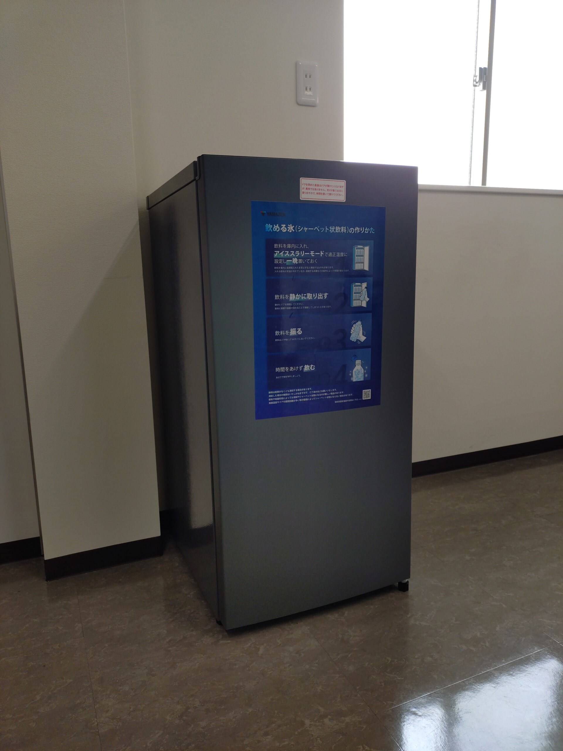 Gray vending machine with a blue poster on its front, located in a hallway near a bright window to the right.