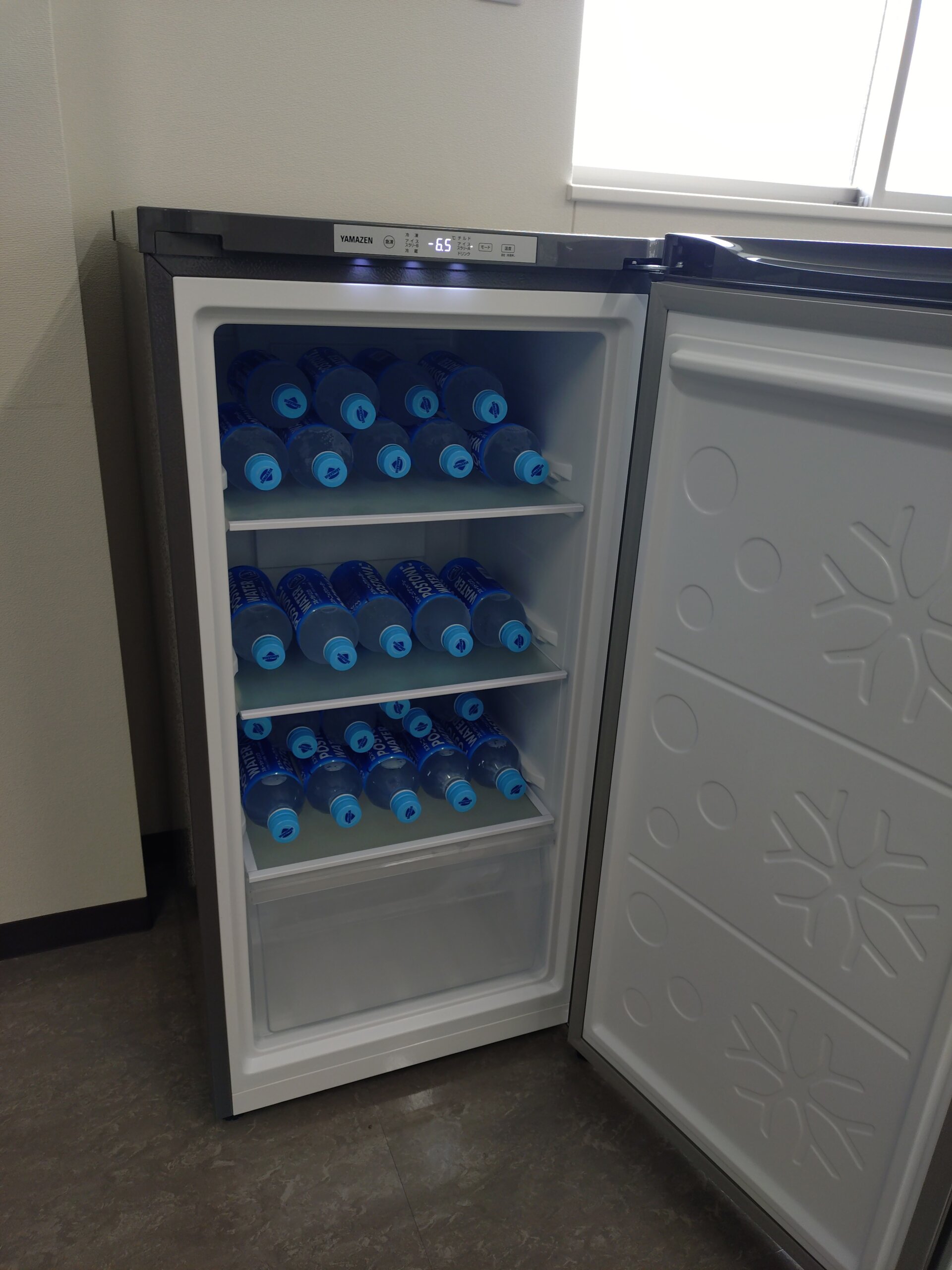 Open small refrigerator filled with bottles of water with blue caps on several shelves inside the unit.